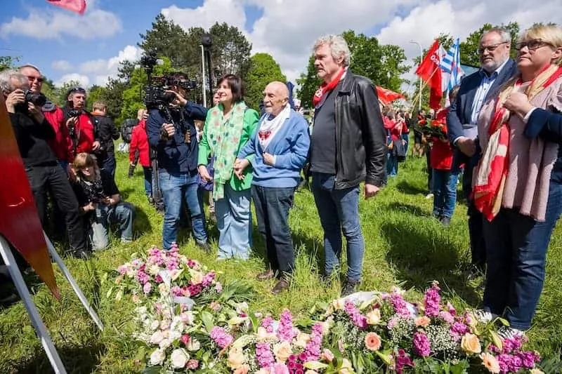Group of people at a memorial event, flowers and flags present.