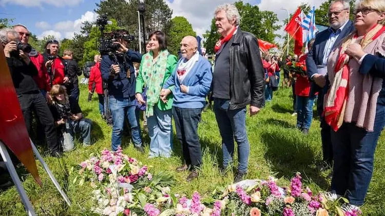 Group of people at a memorial event, flowers and flags present.